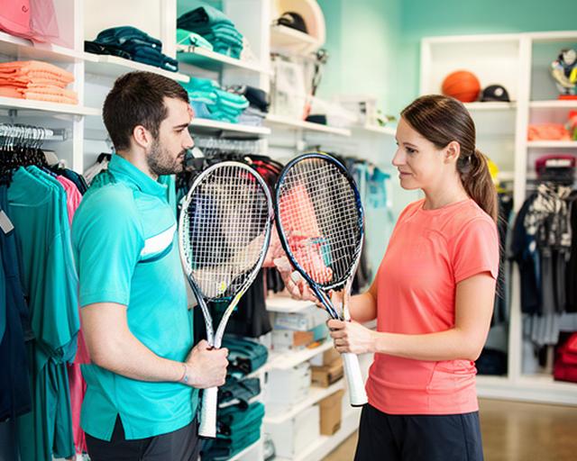 Sports store concierge helping a customer compare tennis rackets beside neatly displayed apparel